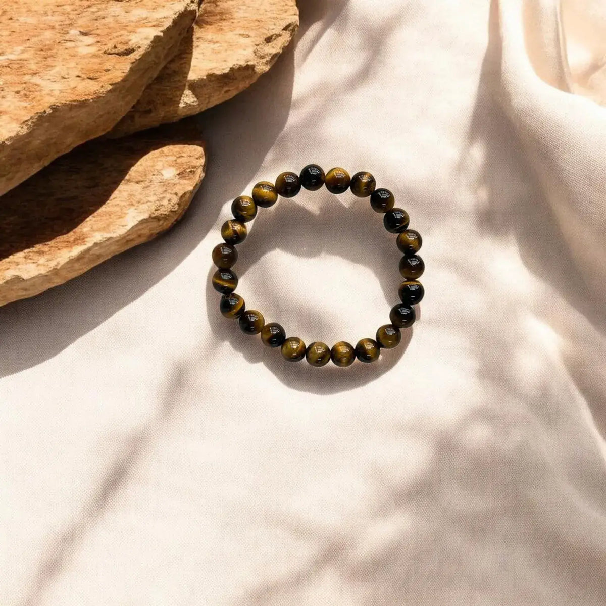 A beaded stretch bracelet made of round tiger's eye beads, styled on a draped cream fabric next to natural stone blocks with soft leaf shadows.