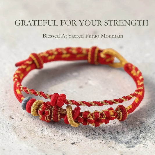 Red and gold bracelet on a textured surface with text 'Grateful for your strength' and 'Blessed at Sacred Putuo Mountain'.