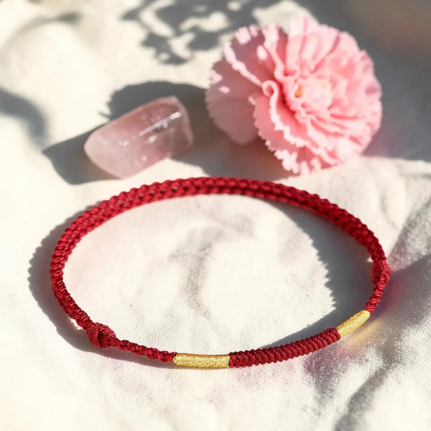 Red braided bracelet with a gold accent on a white surface with a pink flower and clear object.