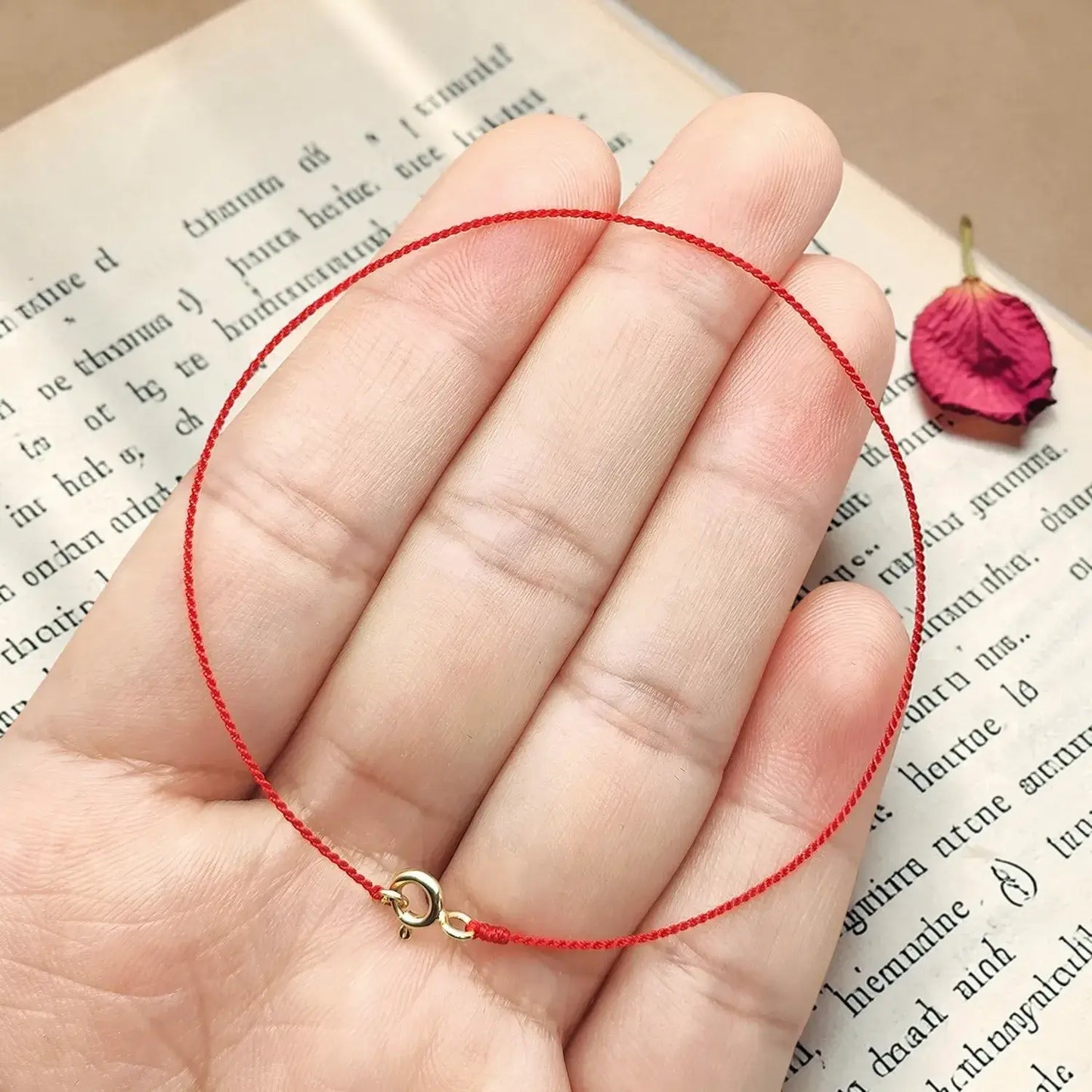 Hand holding a red string bracelet with a gold clasp on an open book with a dried rose petal.
