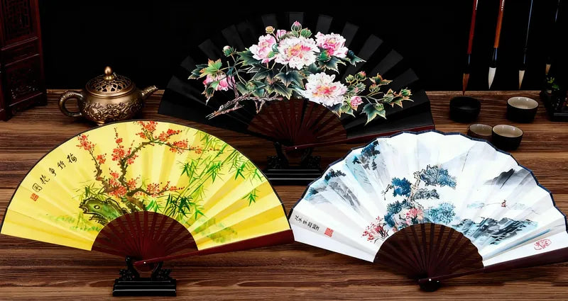 Three decorative fans with floral and landscape designs on a wooden surface.