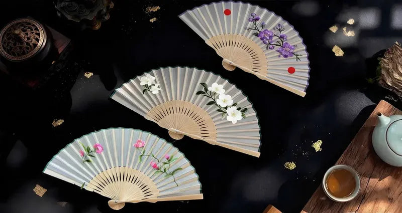 Three decorative fans with floral designs on a dark surface with tea and tea accessories.