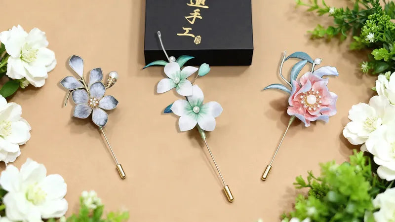 Three floral brooches on a beige surface with greenery and white flowers.