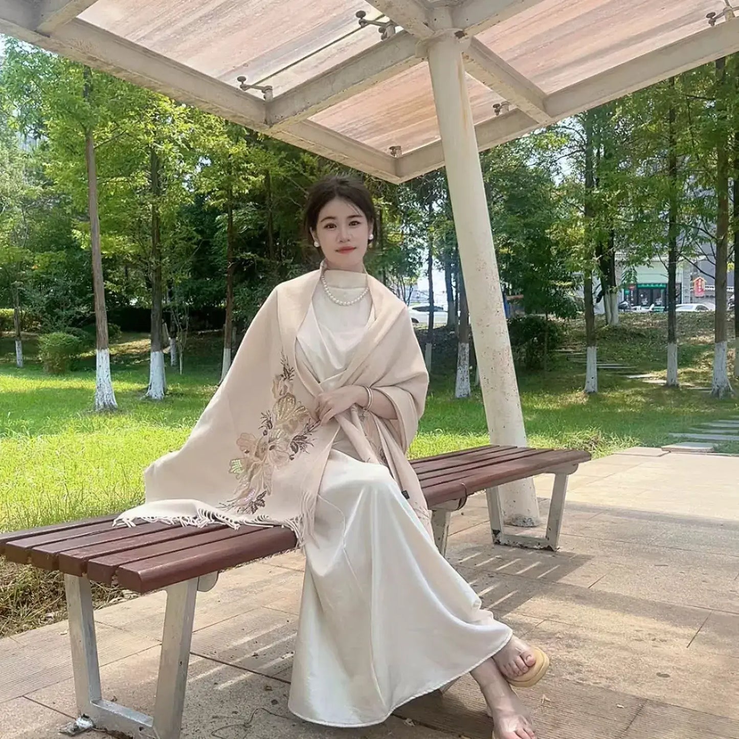 A woman sits on a wooden park bench under a pavilion, wearing a cream-colored satin dress with a beige floral shawl draped over her shoulders.