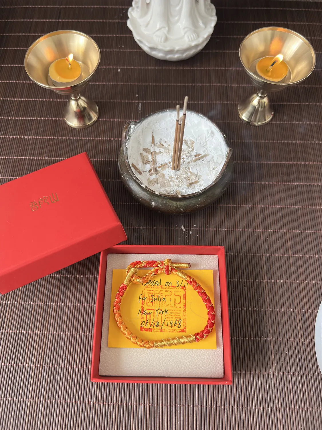 Bracelet with red beads and yellow card in a red box, surrounded by candles and a statue on a textured surface.
