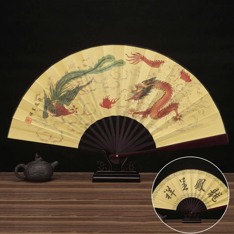 Decorative fan with dragon and phoenix design on a wooden surface with a teapot.
