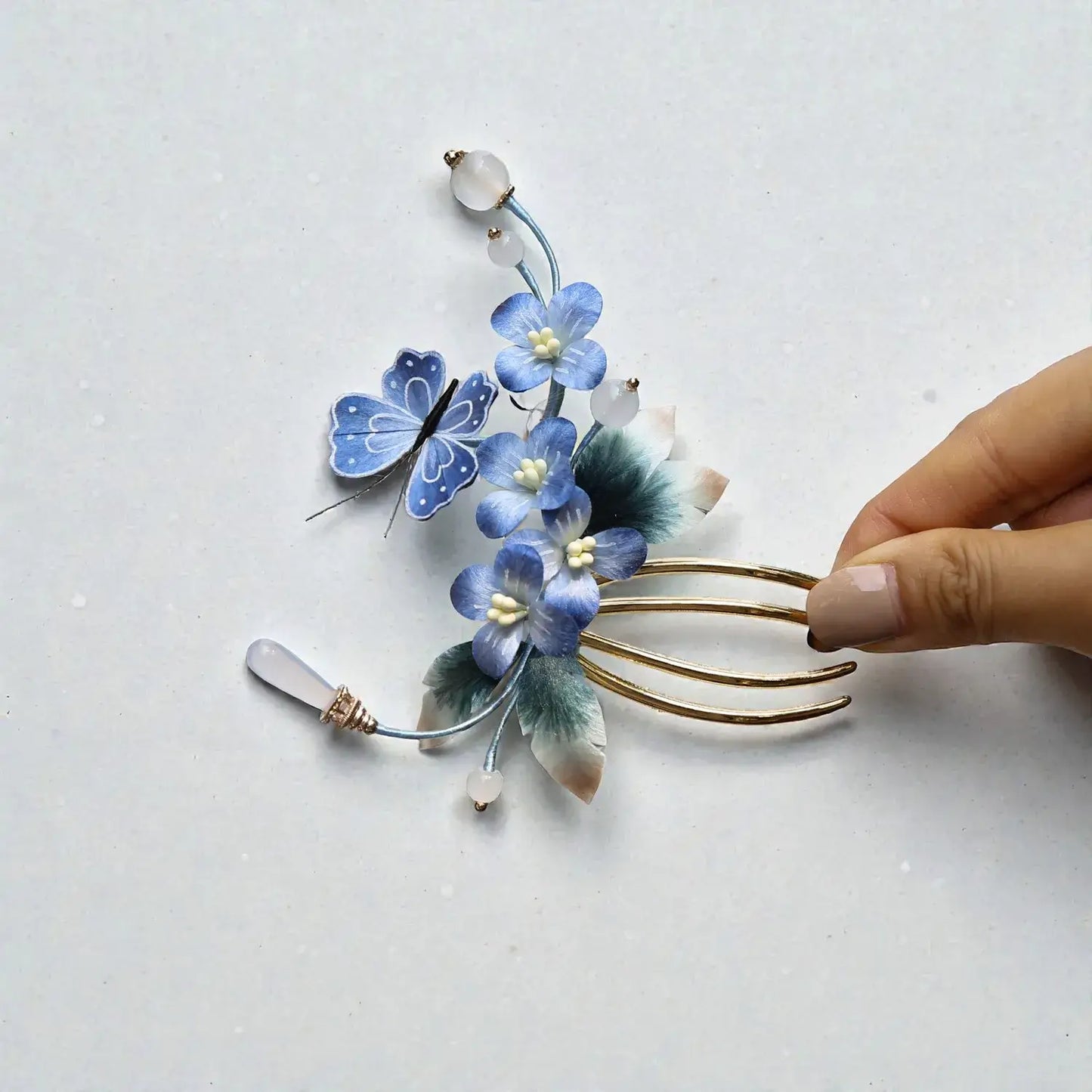 Hand holding a decorative hair comb with blue flowers and a butterfly on a light gray background