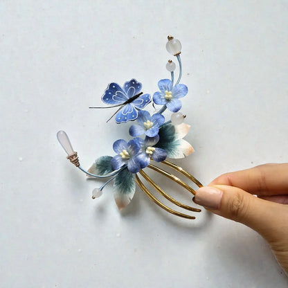 Decorative hair comb with blue flowers and gold accents held by a hand on a light gray background