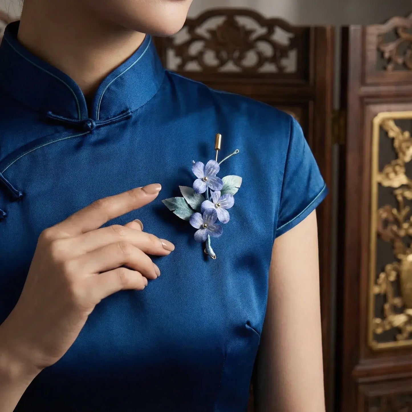 Person wearing a blue dress with a floral brooch, standing in front of an ornate wooden panel.
