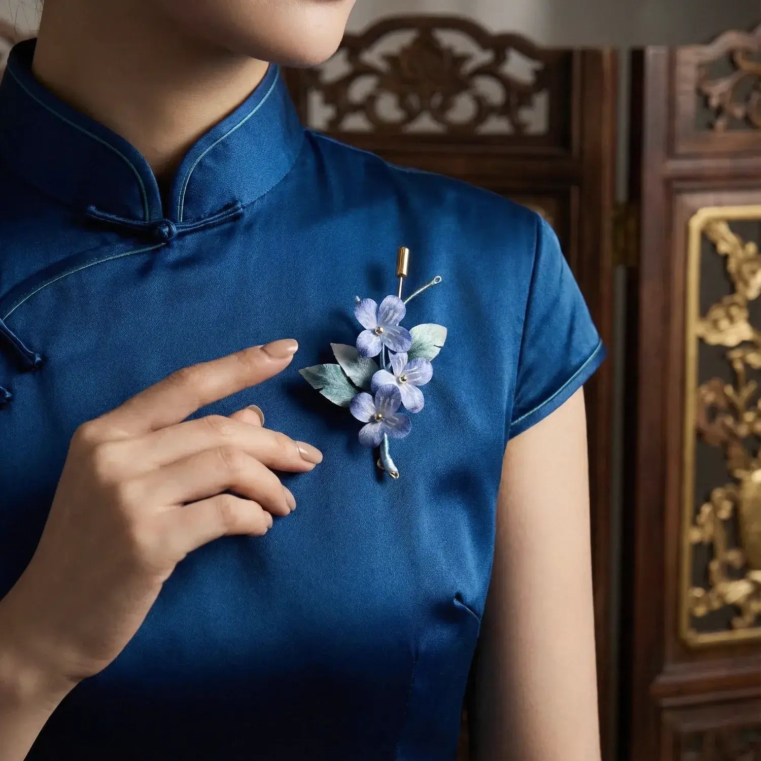 Person wearing a blue dress with a floral brooch, standing in front of an ornate wooden panel.