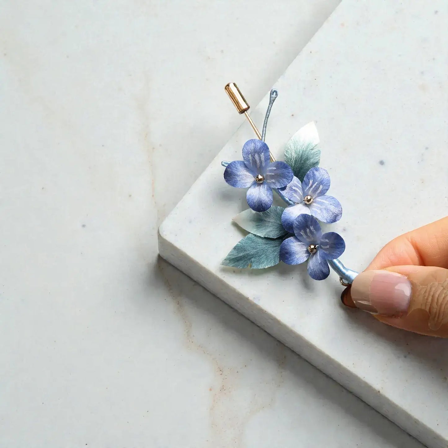 Blue floral hair clip on a marble surface with a hand holding it.