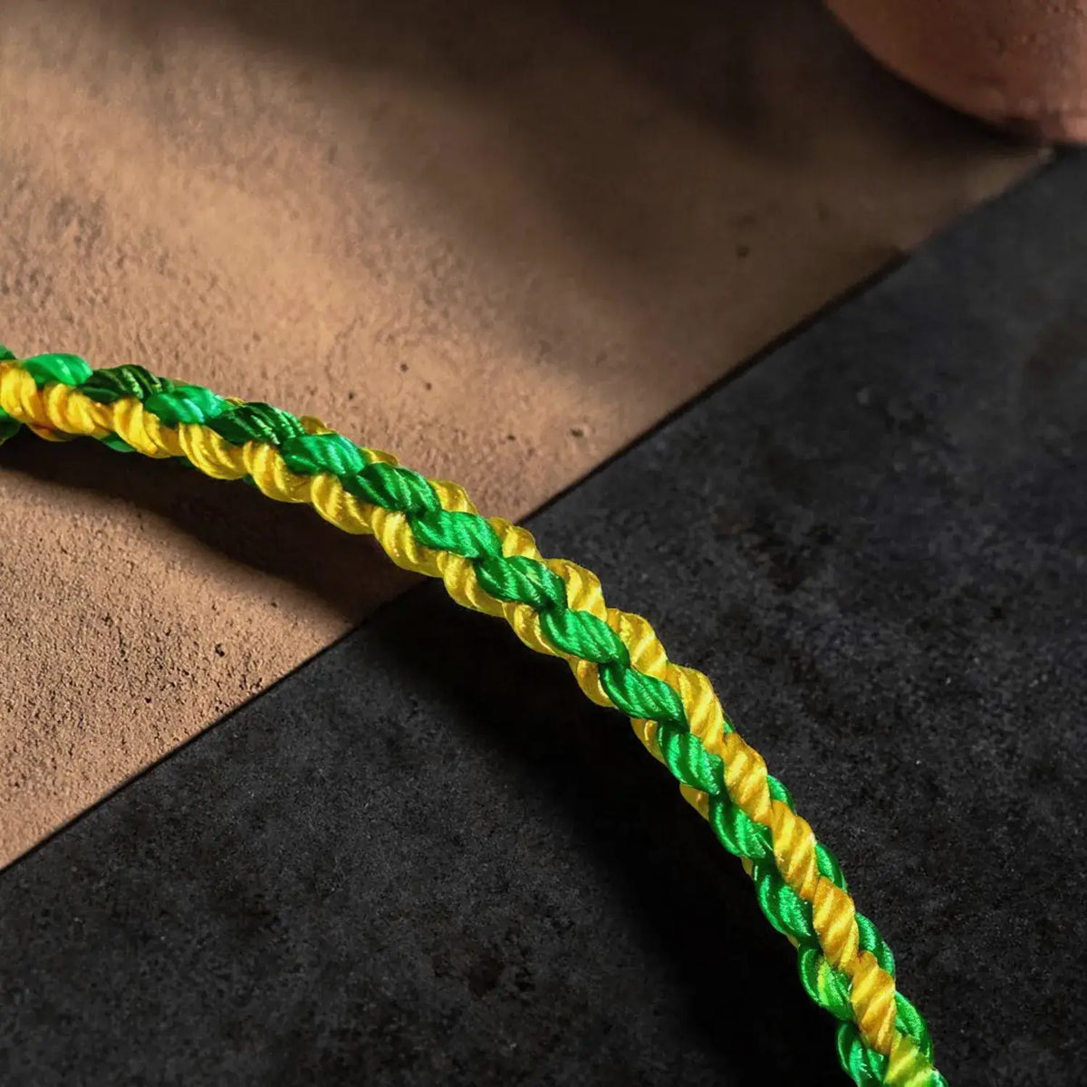 A close-up of a braided rope bracelet in alternating bright green and yellow strands, resting diagonally across a split background of textured beige sand and dark gray stone.