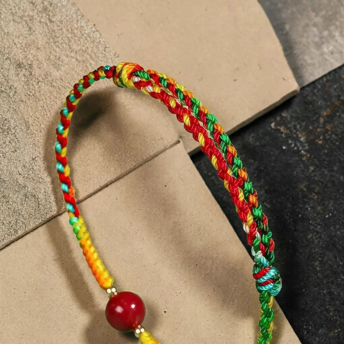 A braided cord bracelet with red, yellow, green, and turquoise strands, featuring a red bead with gold spacer rings, displayed on a textured brown surface.