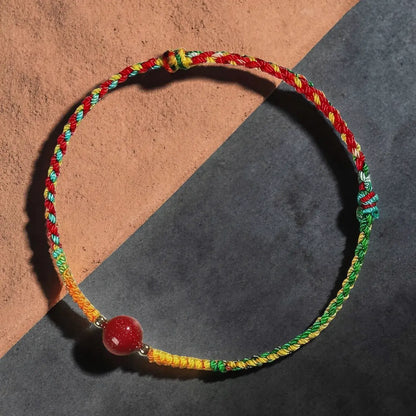 A multicolor braided cord bracelet with a red stone bead, displayed on a split terracotta and dark gray background.
