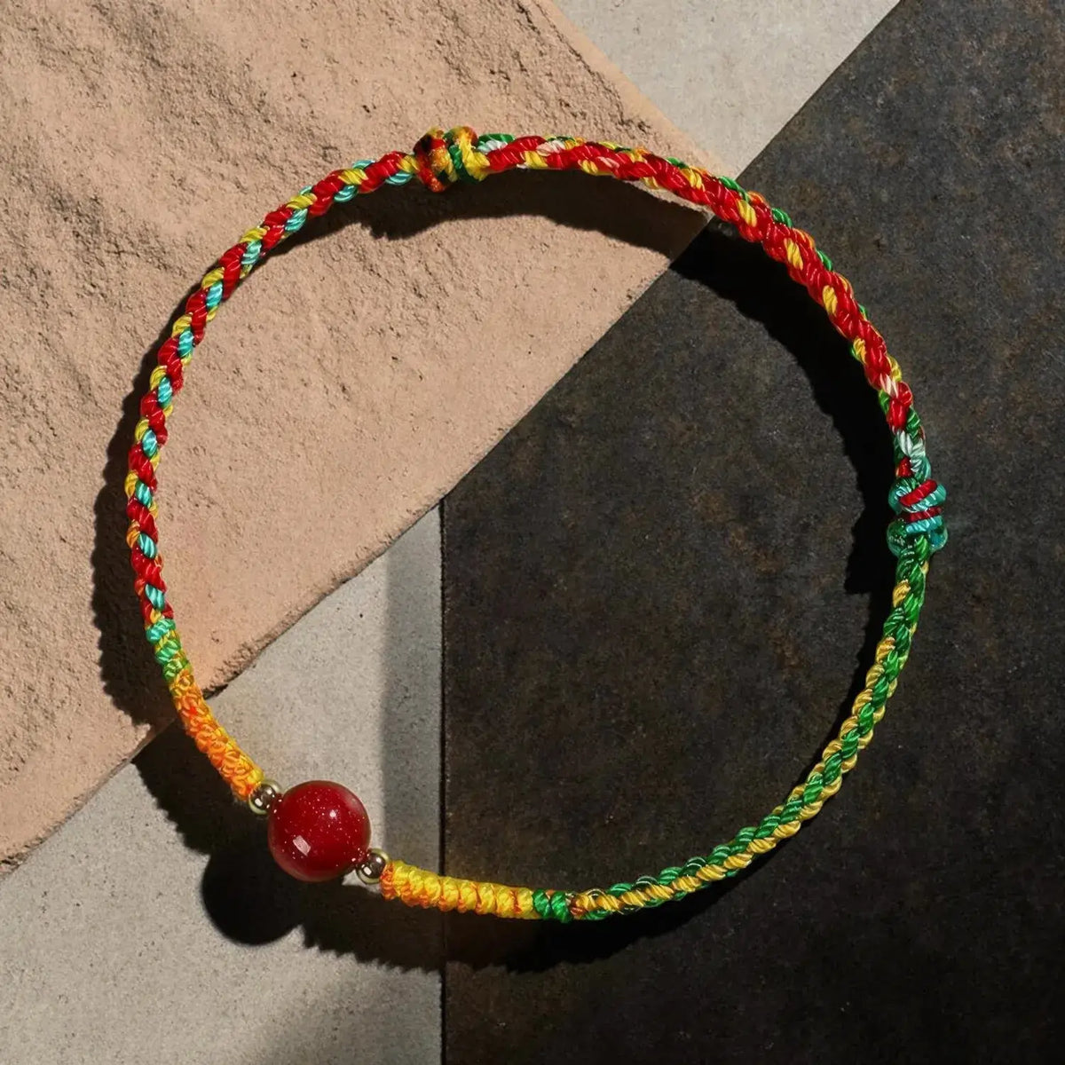 A braided cord bracelet with red, yellow, and green strands, featuring a single red bead and small metallic spacer beads, displayed on a split beige and dark gray background.