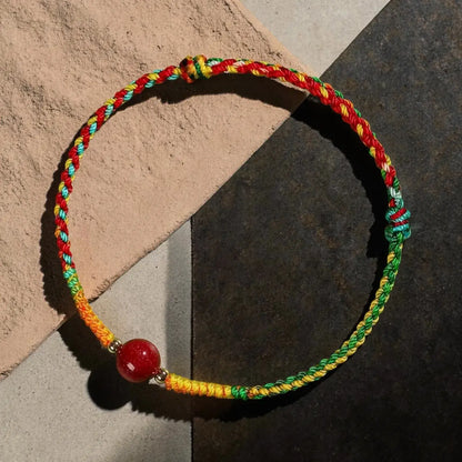 A braided cord bracelet with red, yellow, and green strands, featuring a single red bead and small metallic spacer beads, displayed on a split beige and dark gray background.