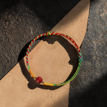 A multicolor braided cord bracelet with a red bead, resting on a split background of dark textured stone and tan paper.