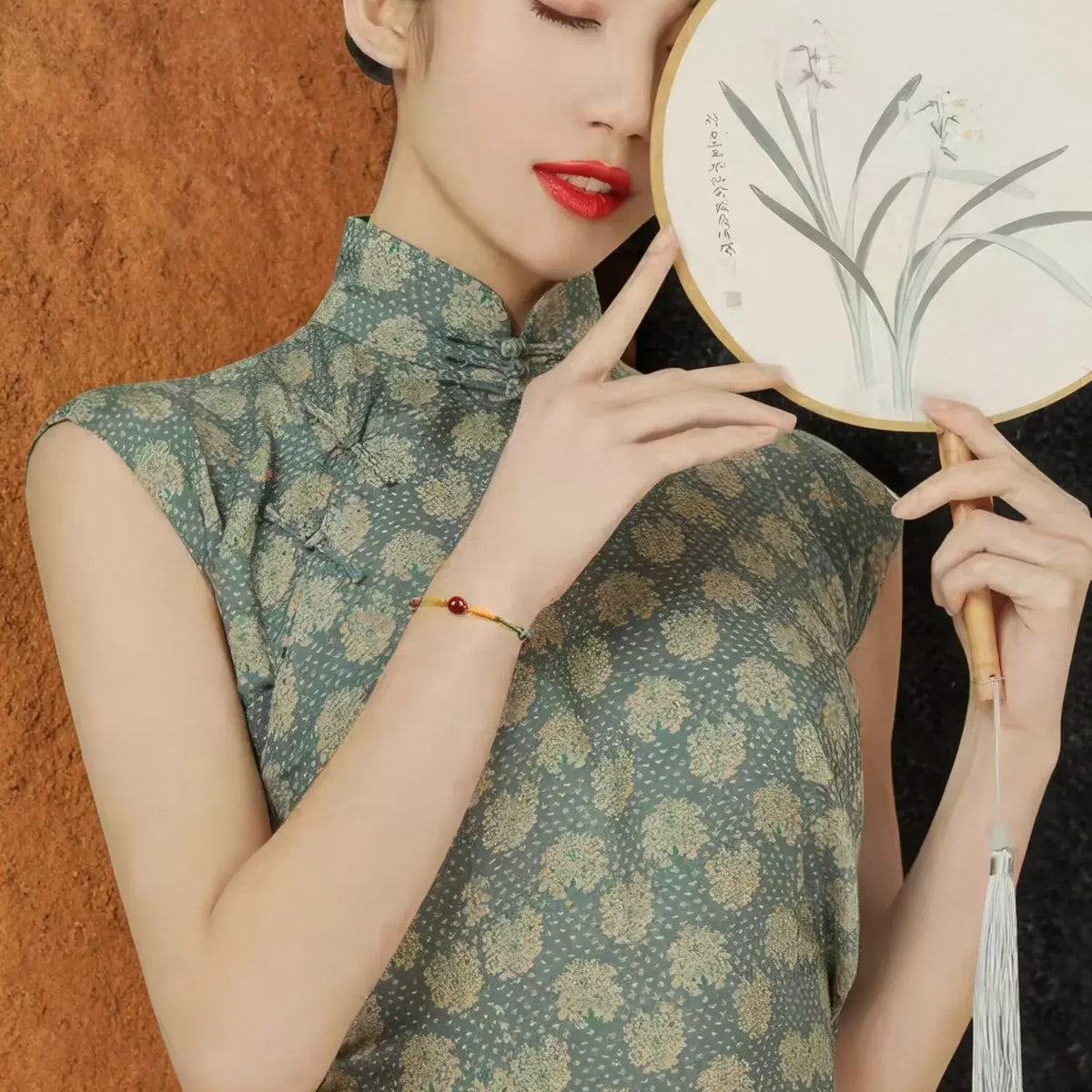 A woman wearing a sleeveless green and beige floral-patterned cheongsam/qipao dress, holding a decorative white fan with a wooden handle. She is wearing a small beaded bracelet on her wrist, posed against a textured brown wall.