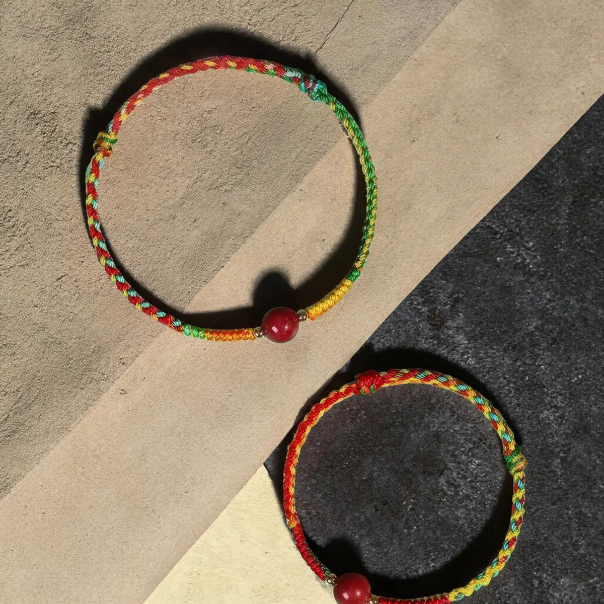 Two braided cord bracelets with red beads, displayed on a split beige and dark gray background.