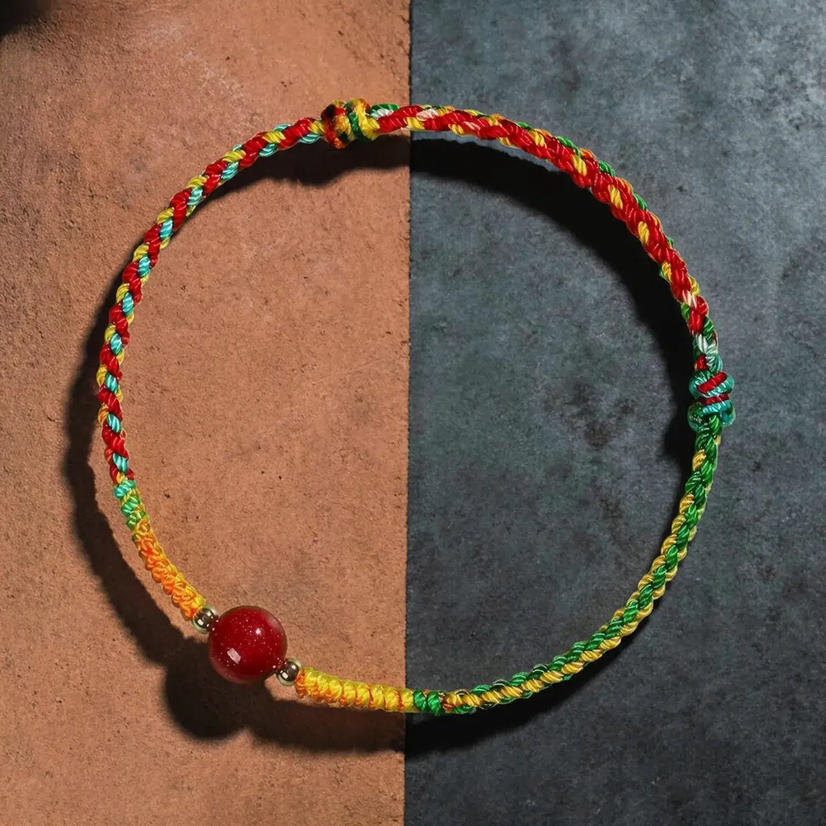 A multicolor braided cord bracelet with a red bead and small metallic spacer, displayed on a split brown and dark gray background.