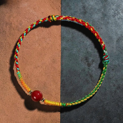 A multicolor braided cord bracelet with a red bead and small metallic spacer, displayed on a split brown and dark gray background.