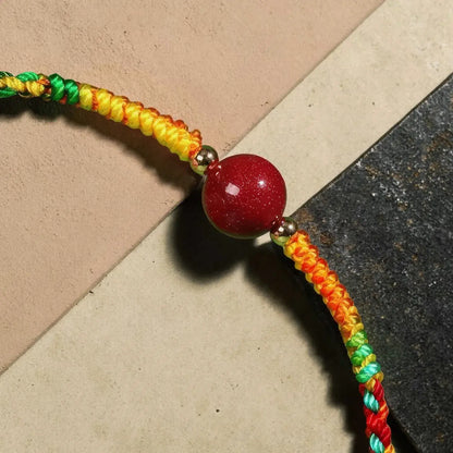 A close-up of a handmade braided cord bracelet with a large red bead at the center, resting on a split beige and dark gray surface.