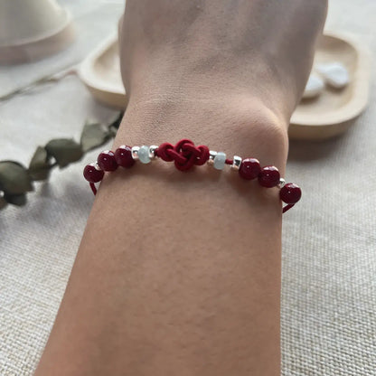 A close-up of a red beaded bracelet with silver-toned spacer beads worn on a person's wrist, set against a soft neutral background with blurred decorative elements.