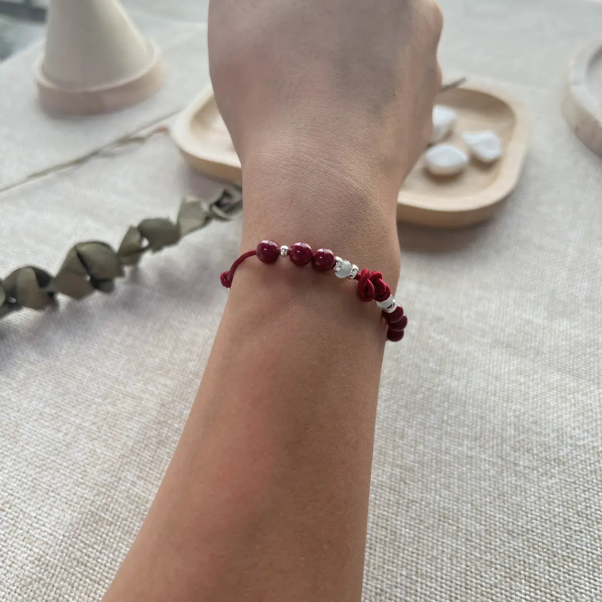 A close-up of a person's wrist wearing a beaded bracelet with deep red beads, a small white bead, and a red-and-white patterned charm, set against a neutral textured fabric background with soft decorative props.
