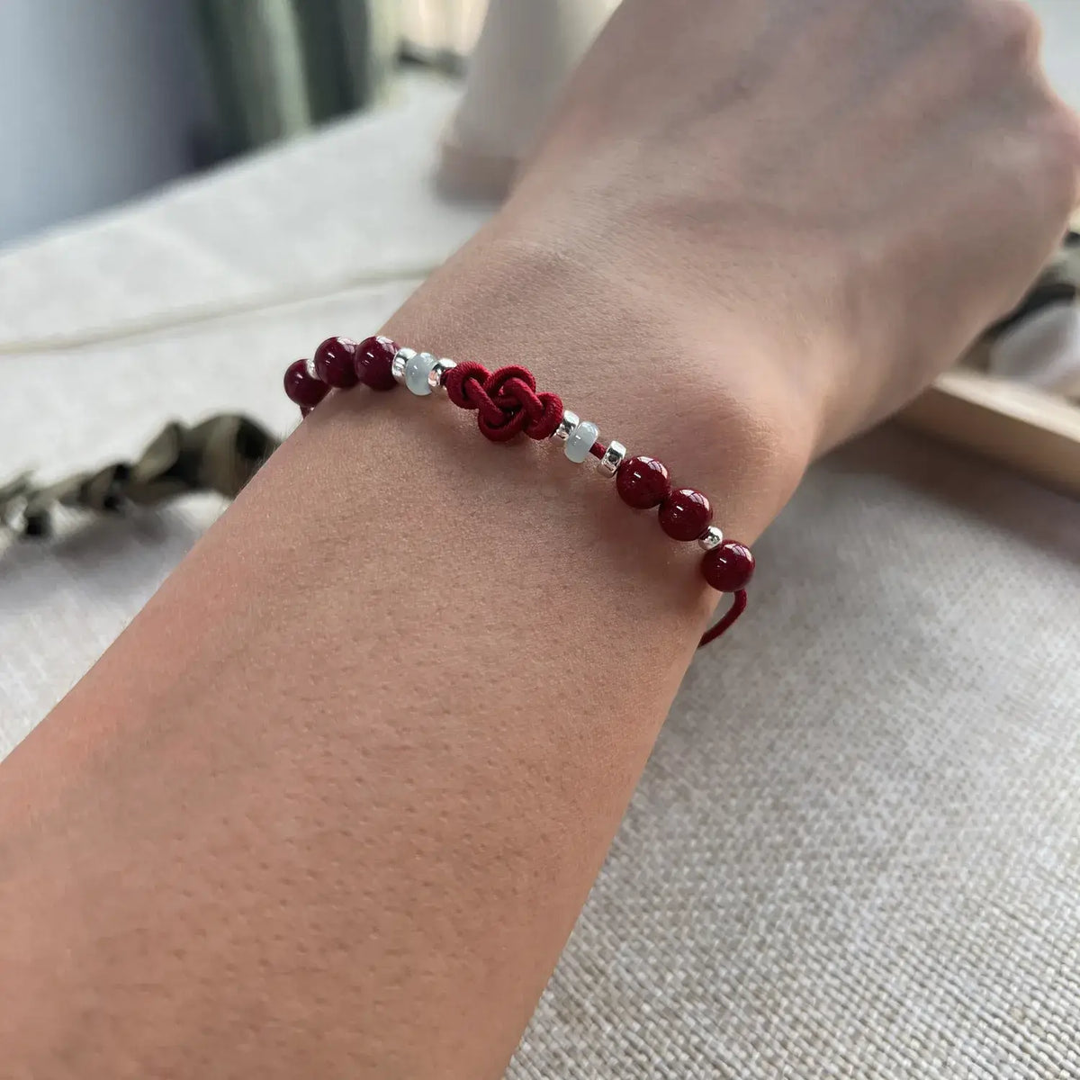A close-up of a person's wrist wearing a red beaded bracelet with a central knot and small silver spacer beads, resting on a light fabric surface.