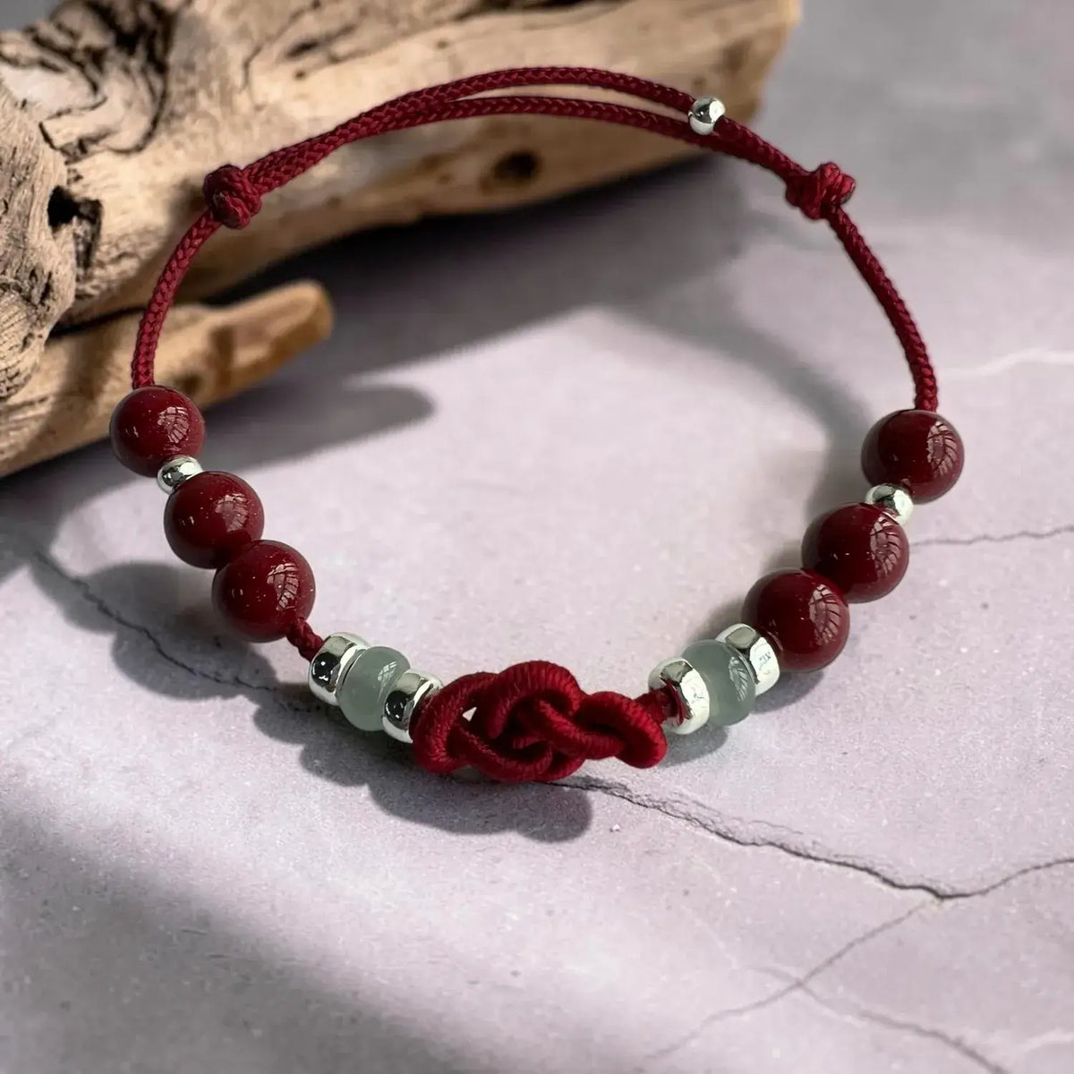 A red cord bracelet with polished dark red beads, light green jade-like spacer beads, and silver metal caps, displayed on a textured gray surface with a piece of driftwood in the background.