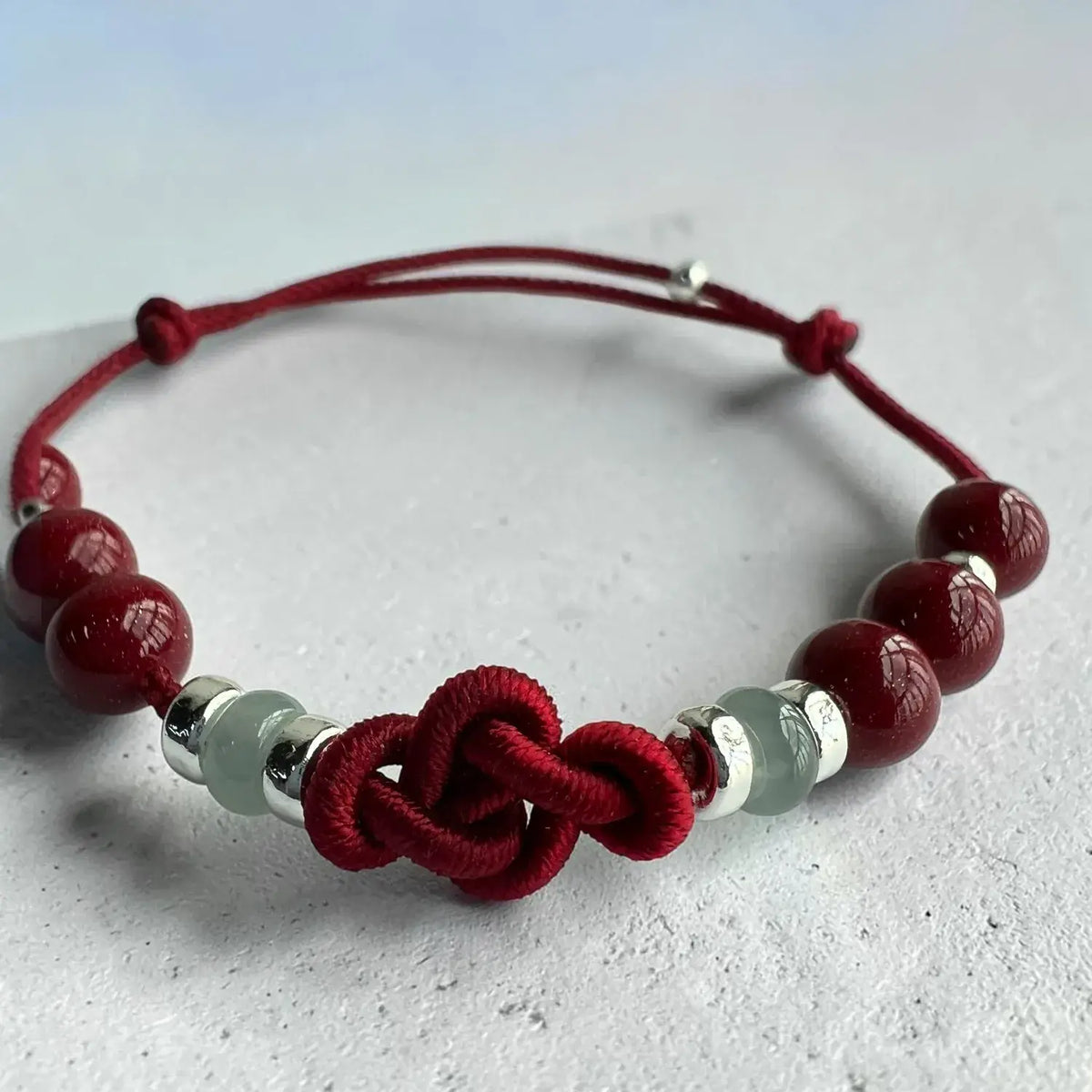 A red cord bracelet with a central knotted motif, accented by dark red round beads and translucent pale green beads separated by shiny silver spacer rings, shown on a light gray textured surface.