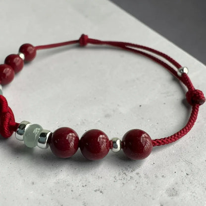 A red cord bracelet with polished dark red round beads, small silver spacer beads, and one pale green translucent bead, displayed on a light gray textured surface.