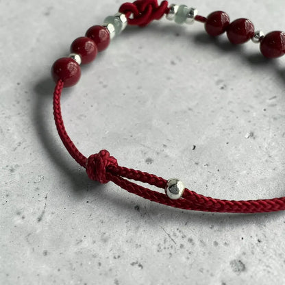 A red braided cord bracelet with glossy red beads, small silver spacer beads, and a knot closure, photographed on a light gray textured surface.
