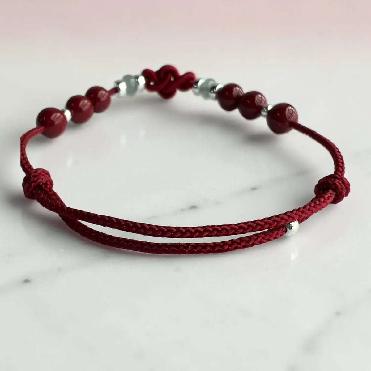 A red braided cord bracelet with dark red round beads and small silver spacer beads, displayed on a light marble surface.