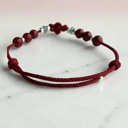A red braided cord bracelet with dark red round beads and small silver spacer beads, displayed on a light marble surface.