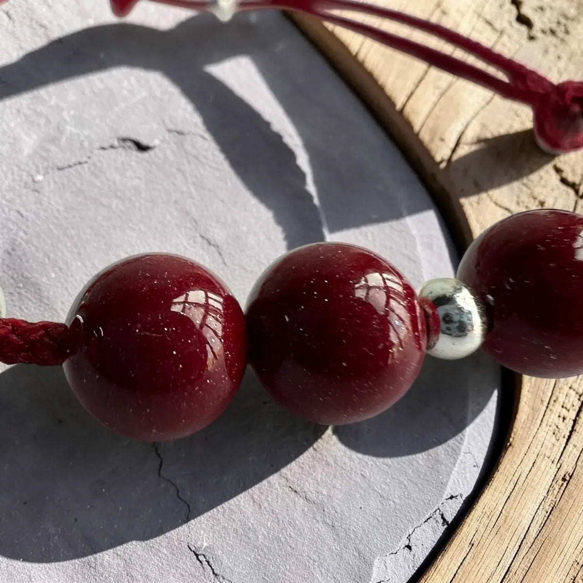 A close-up of a beaded cord accessory with glossy deep red round beads and a small silver spacer, photographed on a gray stone surface with wood visible in the background.