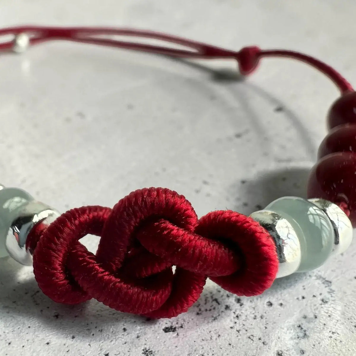 A close-up of a red cord bracelet featuring a knotted central motif, light green translucent beads, and polished silver metal spacers on a textured light gray surface.