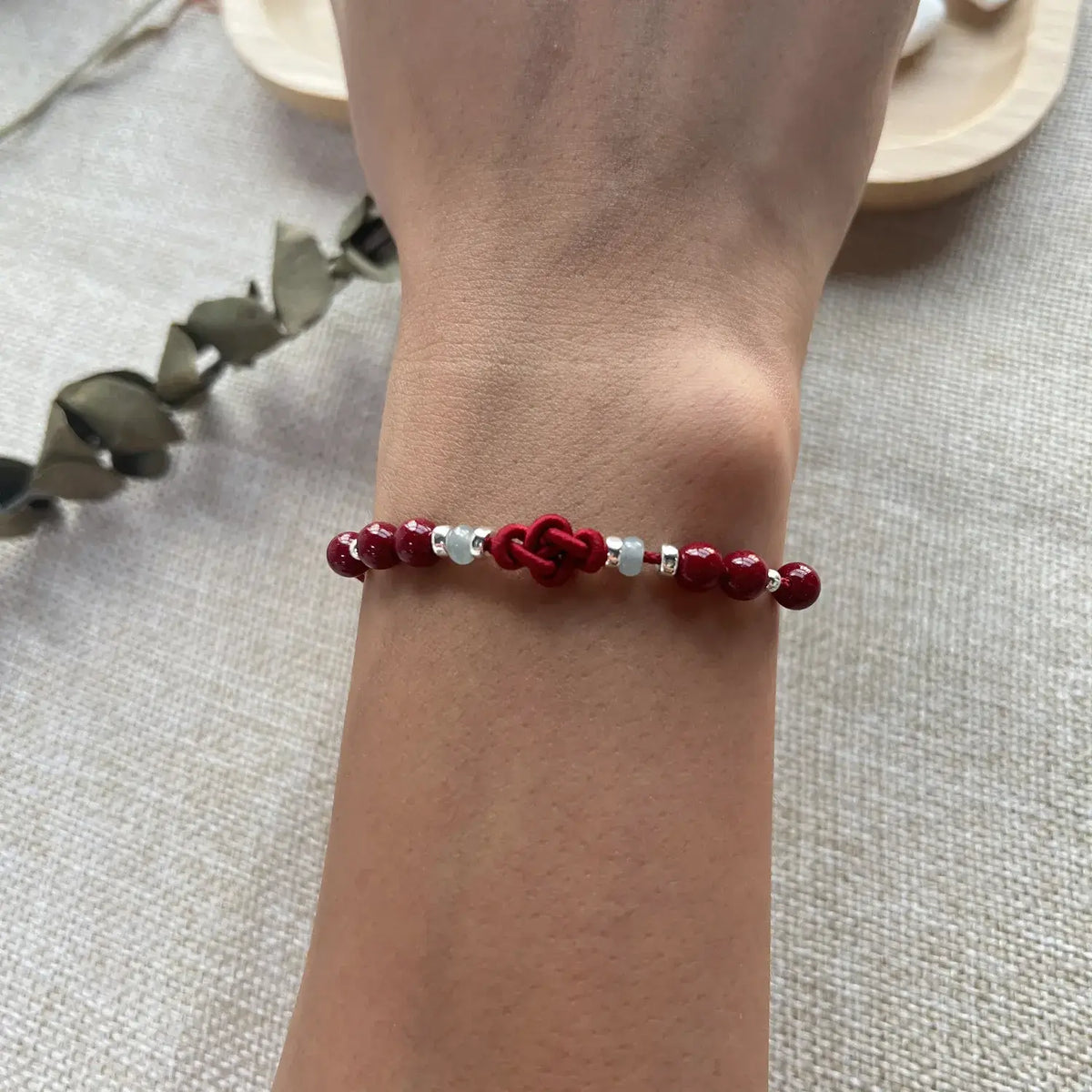 A close-up of a person's wrist wearing a beaded bracelet with dark red round beads, a red knotted center, and small silver spacer beads, set against a neutral textured fabric background.
