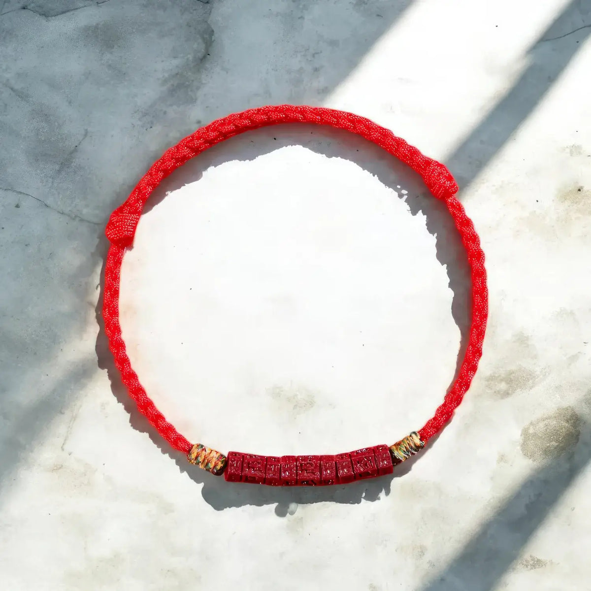 A red braided cord bracelet with a textured red central section and small gold-toned spacer beads, laid flat on a light stone surface with diagonal shadows.