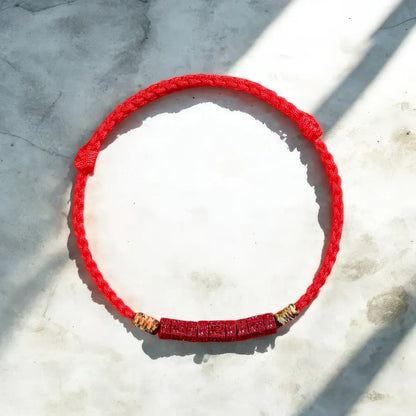 A red braided cord bracelet with a textured red central section and small gold-toned spacer beads, laid flat on a light stone surface with diagonal shadows.