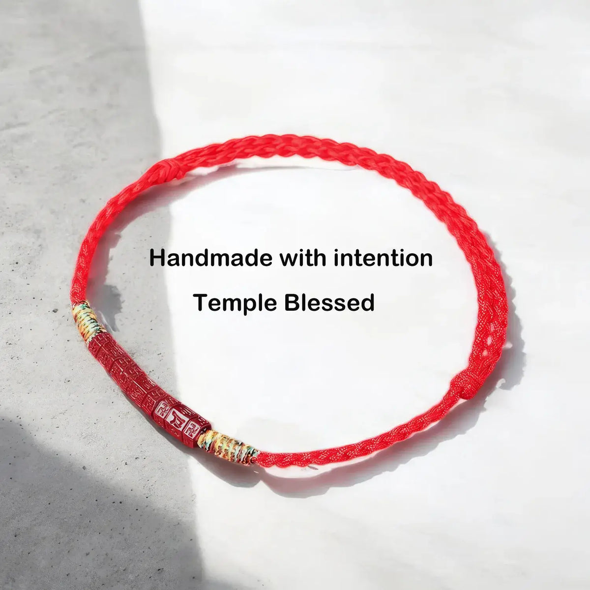 A red braided cord bracelet with gold and silver metallic beads, displayed on a light gray textured surface with a soft shadow. Black text overlay reads 'Handmade with intention Temple Blessed'.