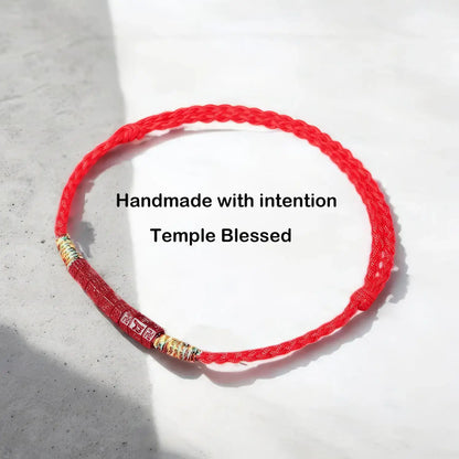 A red braided cord bracelet with gold and silver metallic beads, displayed on a light gray textured surface with a soft shadow. Black text overlay reads 'Handmade with intention Temple Blessed'.