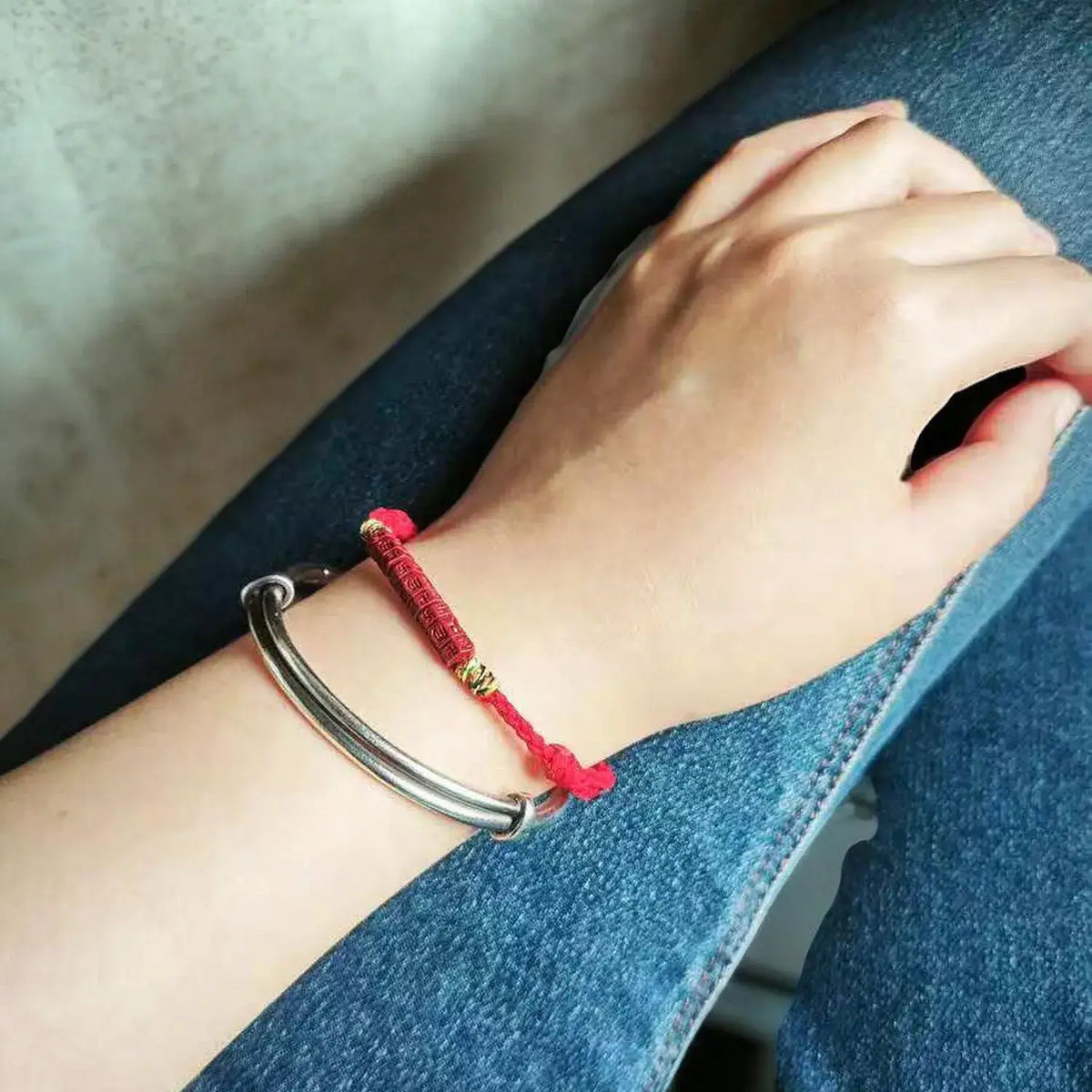 A close-up of a person's wrist wearing two bracelets: a silver-toned metal cuff bracelet and a red cord bracelet with a small gold bead accent, resting on blue denim.