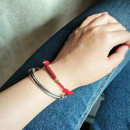 A close-up of a person's wrist wearing two bracelets: a silver-toned metal cuff bracelet and a red cord bracelet with a small gold bead accent, resting on blue denim.