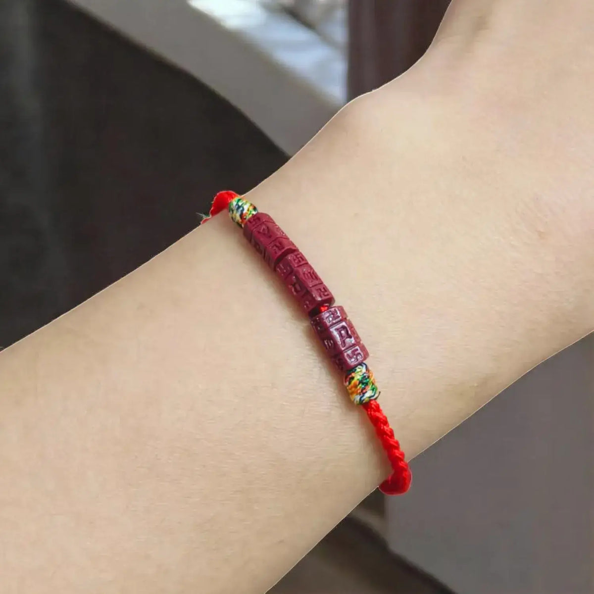 A red Chinese-style cord bracelet with carved red beads and small gold and green decorative accents, shown worn on a person's wrist.