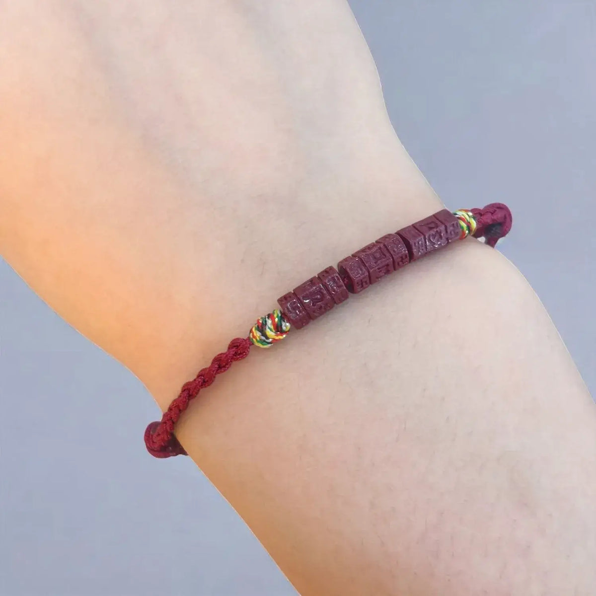 A dark red beaded bracelet with Tibetan script carvings and small multicolor accent beads, shown worn on a person's wrist against a soft gray background.