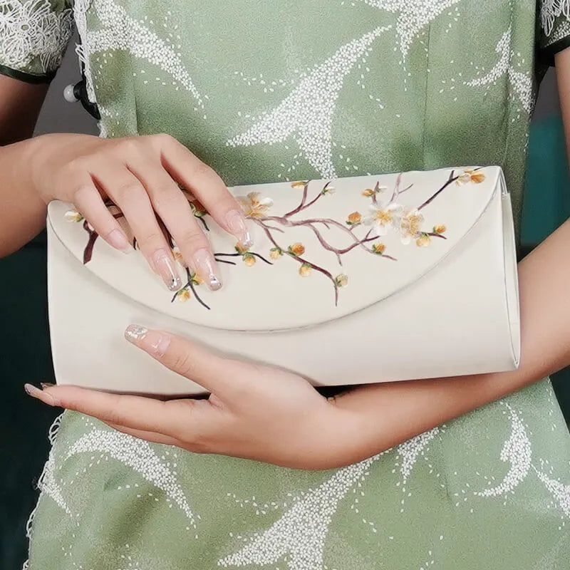 Hand holding a white clutch with floral design, person wearing a green dress with floral pattern.