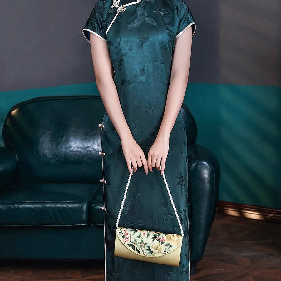 Woman in a dark green traditional dress holding a matching handbag in a room with a blue wall and black leather couch.