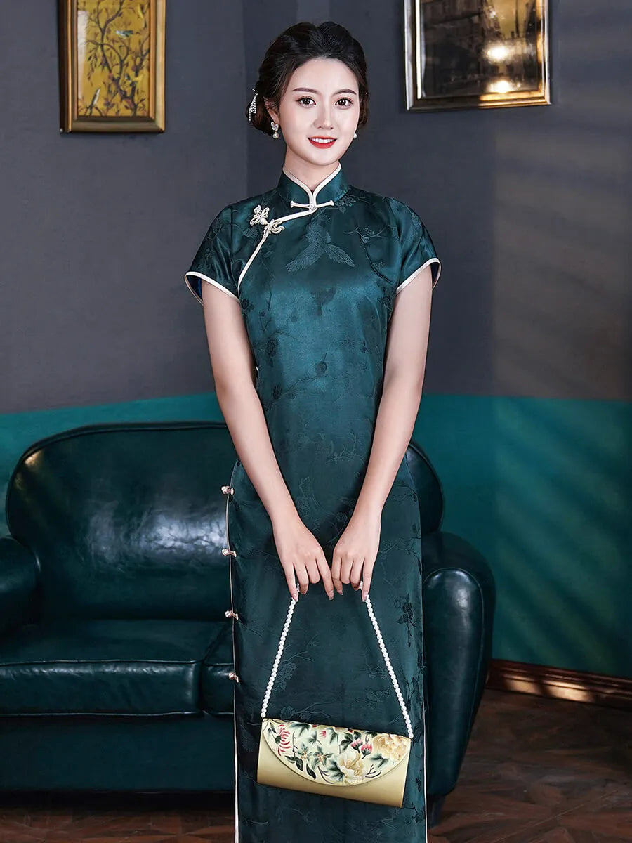 Woman in a dark green traditional dress holding a matching handbag in a room with a blue wall and black leather couch.