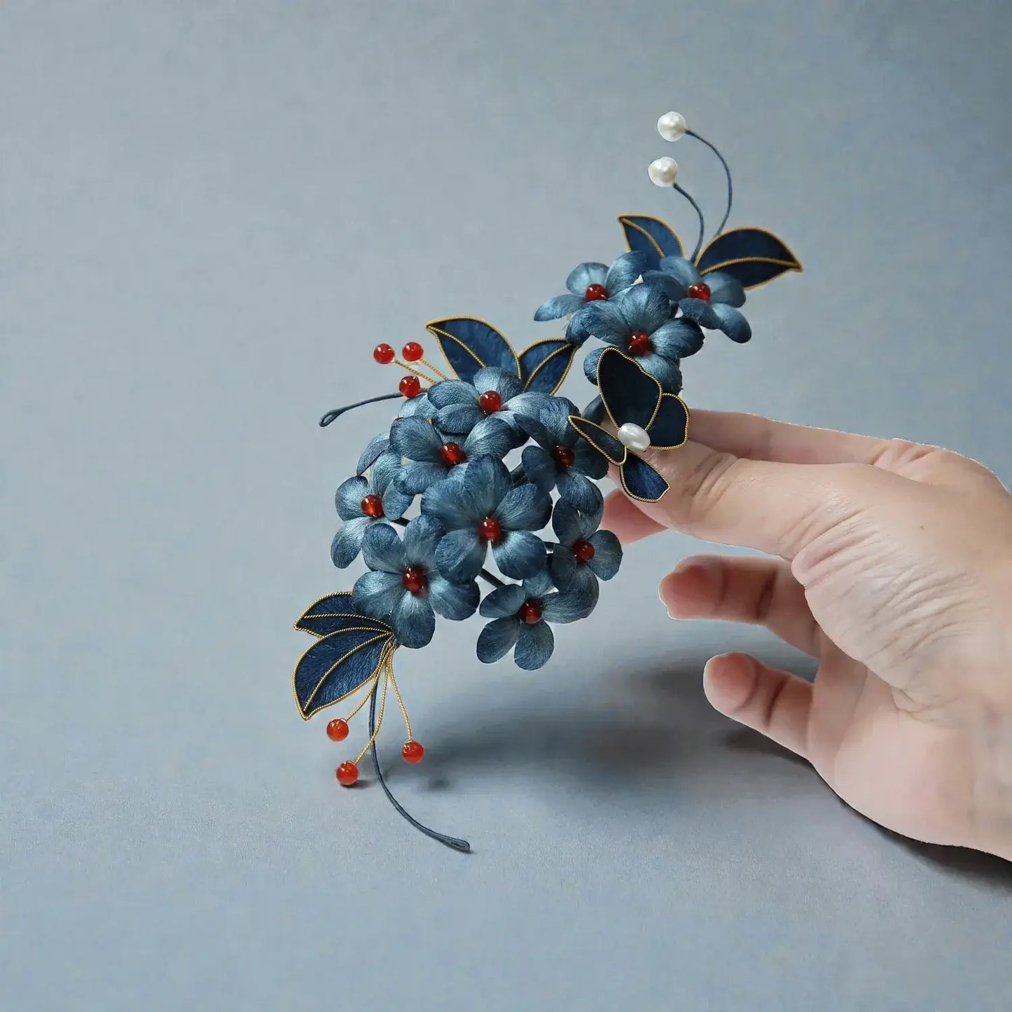Hand holding a decorative hairpiece with blue flowers and red berries against a gray background